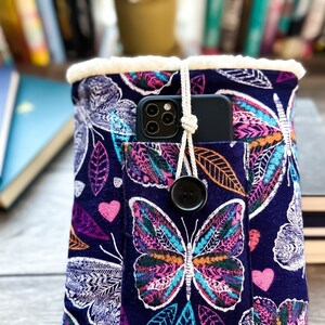 May include: A blue fabric pouch with a white fuzzy lining and a zipper closure. The pouch has a pocket on the front with a phone inside. The pouch is decorated with a colorful butterfly pattern.