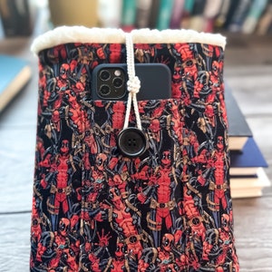 Op de afbeelding: Een zwarte en rode stoffen tas met een witte fleece voering. De tas heeft een herhalend patroon van Deadpool-karakters. Een zwarte telefoon met een wit snoer zit in de zak van de tas.