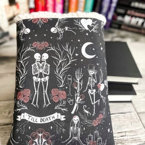 May include: Black book cover with a white and pink floral pattern featuring skeletons, hearts, and the text "Till Death".