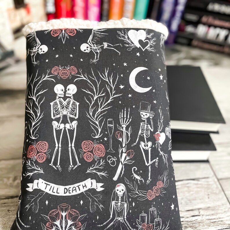 May include: Black book cover with a white and pink floral pattern featuring skeletons, hearts, and the text "Till Death".
