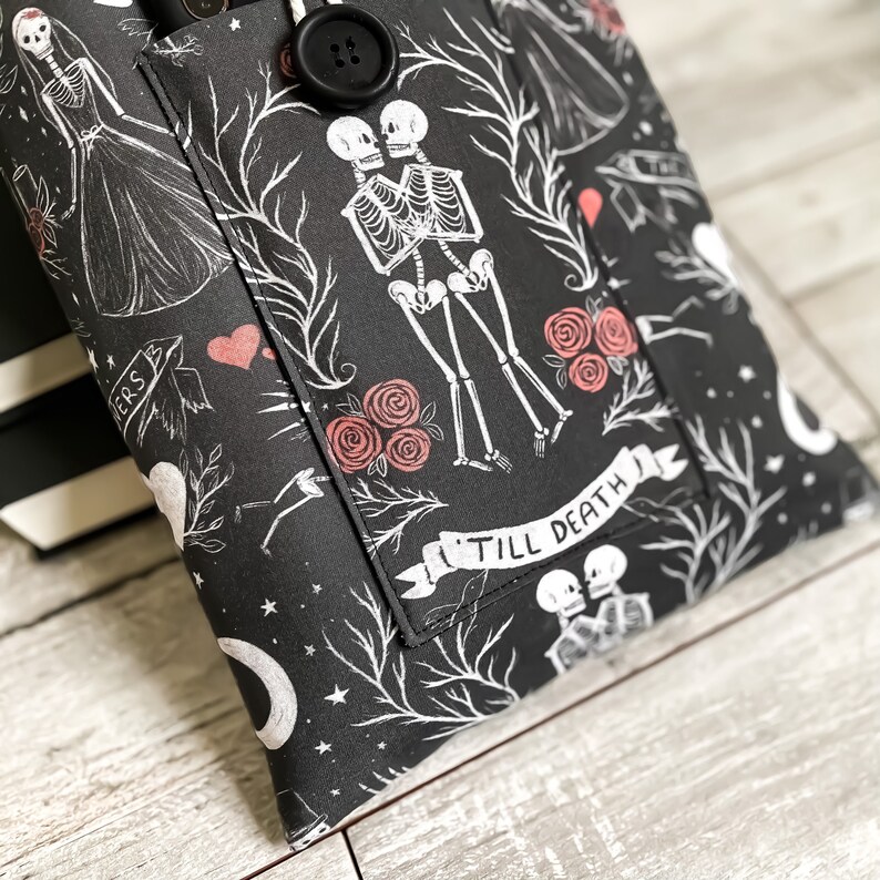 May include: Black fabric tote bag with a white and red print of skeletons holding hands and roses. The bag has a pocket with a button closure and the text "Till Death" on a banner.