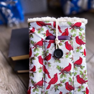 Pode incluir: Uma capa de livro de tecido com um padrão de cardeais vermelhos e folhas de azevinho sobre um fundo cinza claro. A capa tem um forro cremoso e fofo e um bolso com fecho de botão preto. Um cordão creme prende a parte superior.