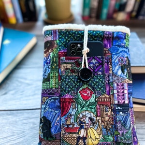 Può includere: Una colorata custodia per libri trapuntata con un design a vetrata raffigurante scene di una fiaba. La custodia ha una tasca, una chiusura a bottone e un bordo color crema. Uno smartphone nero è nella tasca.