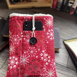 May include: Red fabric book cover with white snowflake pattern and a black button closure. The cover has a pocket holding a black smartphone. The interior is lined with white fluffy material.