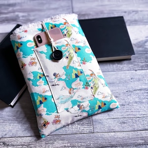 May include: A white and teal fabric book sleeve featuring a map design with Peter Pan characters. The sleeve has a pocket for a phone and a black button closure. The interior is lined with white fluffy fabric.