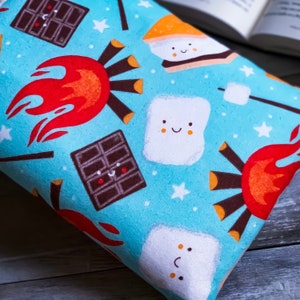 May include: A light blue fabric with a repeating pattern of campfire elements. The design includes smiling marshmallows, chocolate bars, campfires, and s'mores. White stars are scattered across the background. The fabric is folded, with a book visible in the background.