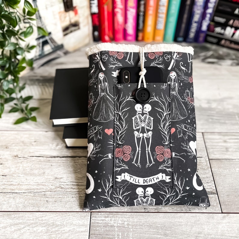 May include: Black and white phone case with a skeleton couple design. The case has a pocket for a phone and a button closure. The text "Till Death" is printed on the case.