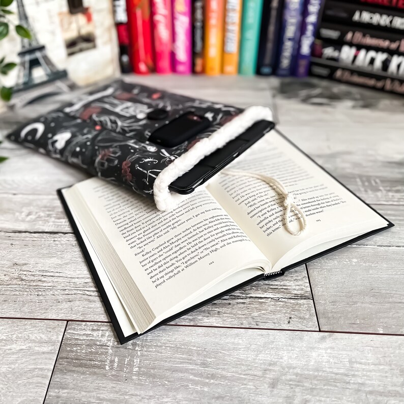 May include: A black and white book sleeve with a white fluffy lining. The sleeve is decorated with a whimsical design of stars, moons, and other celestial elements. The sleeve is holding a black e-reader and a book.