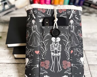Pode incluir: Capa de livro preta com um padrão floral branco e rosa com esqueletos de mãos dadas e o texto "Till Death". A capa tem um bolso para um telefone e um fecho de botão.