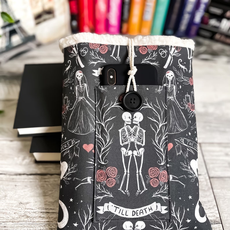 May include: Black book cover with a white and pink floral pattern featuring skeletons holding hands and the text "Till Death". The cover has a pocket for a phone and a button closure.