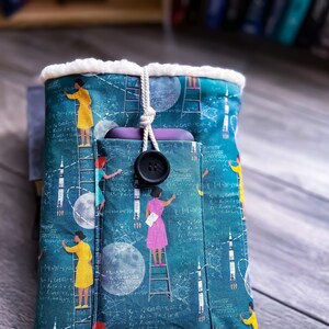 Pode incluir: Uma bolsa de tecido azul-esverdeado com forro de lã branca. A bolsa apresenta um padrão repetitivo de mulheres com vestidos amarelos e rosa escalando escadas, foguetes e luas. A bolsa tem um bolso na frente com um telefone roxo dentro. Um botão preto grande está preso à bolsa.