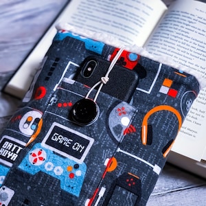 May include: A gray and blue fabric book sleeve with a pocket for a phone and a headphone design. The sleeve has a white fuzzy lining and the text "GAME ON" printed on it.