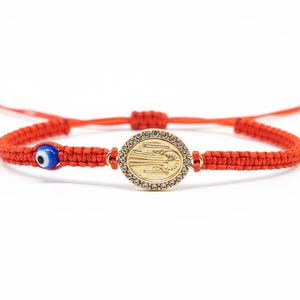 May include: Red macrame bracelet featuring a gold-colored oval pendant with a religious figure and a blue evil eye charm. The pendant is surrounded by small, clear crystals. The bracelet is adjustable.