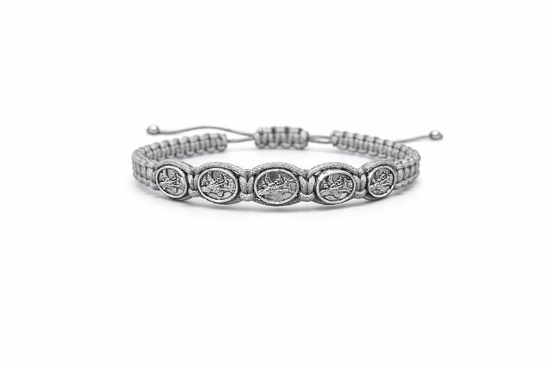 May include: A grey macrame bracelet with five oval, silver-toned medallions. Each medallion has a detailed relief design. The bracelet has an adjustable sliding knot closure. The bracelet is displayed on a white background.