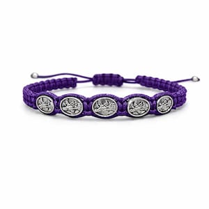 May include: A purple braided bracelet with five silver-coloured oval charms. Each charm depicts a religious scene. The bracelet has adjustable sliding knots. The background is plain white.