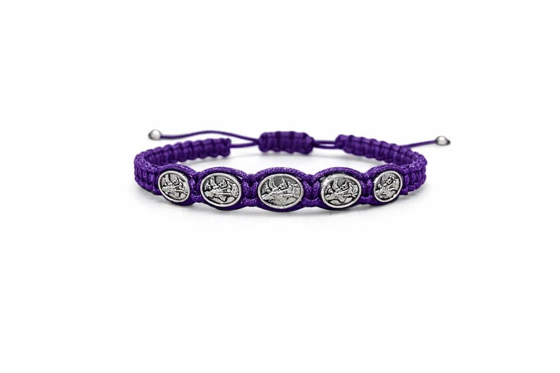 May include: A purple braided bracelet with five silver-coloured oval charms. Each charm depicts a religious scene. The bracelet has adjustable sliding knots. The background is plain white.