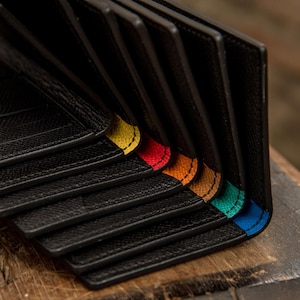 May include: A stack of black leather wallets with colorful stitching along the edges. The stitching colors are yellow, red, orange, teal, and blue.