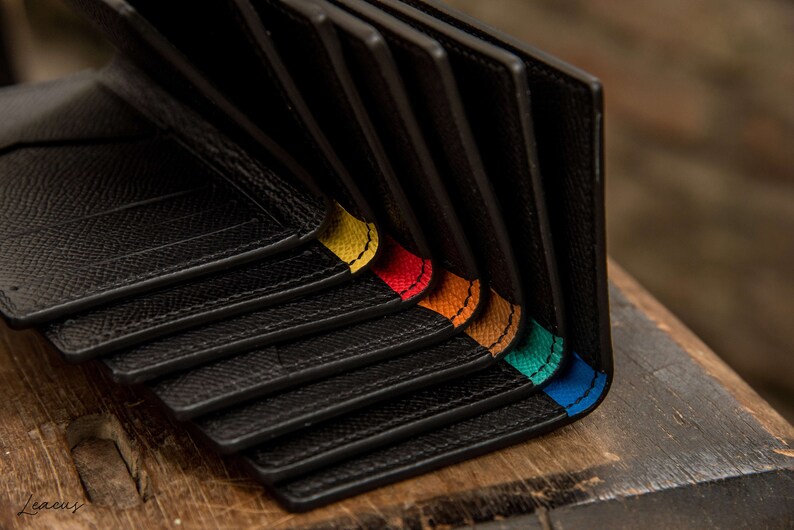 May include: A stack of black leather wallets with colorful stitching along the edges. The stitching colors are yellow, red, orange, teal, and blue.