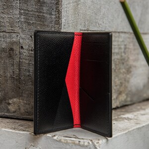 May include: A black leather wallet with a red interior. The wallet is open and the inside is visible. The wallet is on a gray surface.