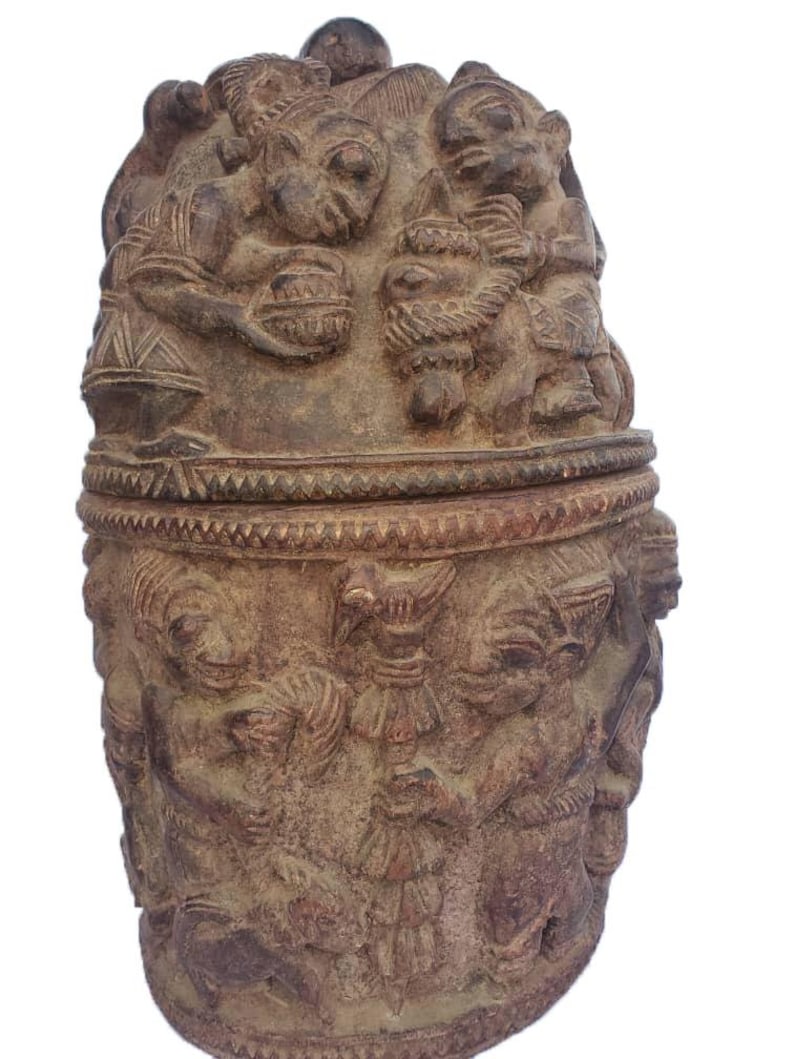May include: A brown, carved wooden container with a lid. The container is covered in detailed relief carvings of figures and patterns. The lid has a small knob on top. The container is likely a decorative or functional item.