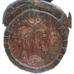 Könnte beinhalten: Ein geschnitztes Holzartefakt mit einem zentralen ovalen Design mit einem Gesicht und anderen Figuren. Der äußere Rahmen ist dunkelbraun mit einem ziegelartigen Muster. Die Oberseite hat ein geschnitztes Design, und die Gesamtfarbe ist eine Mischung aus Braun und Rotbraun.