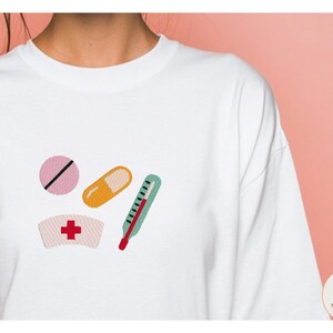 May include: A white long-sleeve shirt featuring an embroidered design of a pink round pill with a black line, an orange capsule, a teal thermometer with a red tip, and a white nurse cap with a red cross symbol centered on the front chest area.