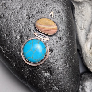 May include: A pendant featuring a circular bright blue stone with a gold-toned crackle pattern set in a copper frame, connected to an oval-shaped stone with horizontal tan, brown, and cream bands, also encased in a dark metal setting.