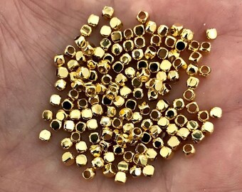 May include: A pile of gold-colored metal beads with a square shape and a hole in the center. The beads are scattered on a hand.