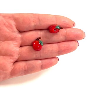 Pode incluir: Duas contas de vidro vermelho em forma de maçã com folhas verdes. As contas têm um diâmetro de aproximadamente 2,5 cm.