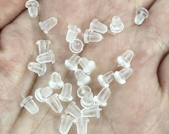 May include: A close-up of a pile of clear plastic earring backs. The backs are small and round, with a small hole in the center. They are designed to be used with stud earrings.