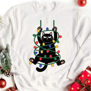 Puede incluir: Sudadera blanca con un gato negro envuelto en luces navideñas. El gato tiene una expresión de enfado y se agarra a líneas verdes. Las luces son rojas, azules, amarillas y verdes. Un diseño festivo para las fiestas.