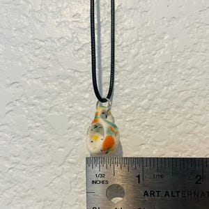 May include: A small, speckled ceramic pendant necklace with a black cord. The pendant is white with orange, green, and yellow accents. The pendant measures approximately 0.75 inches in height.