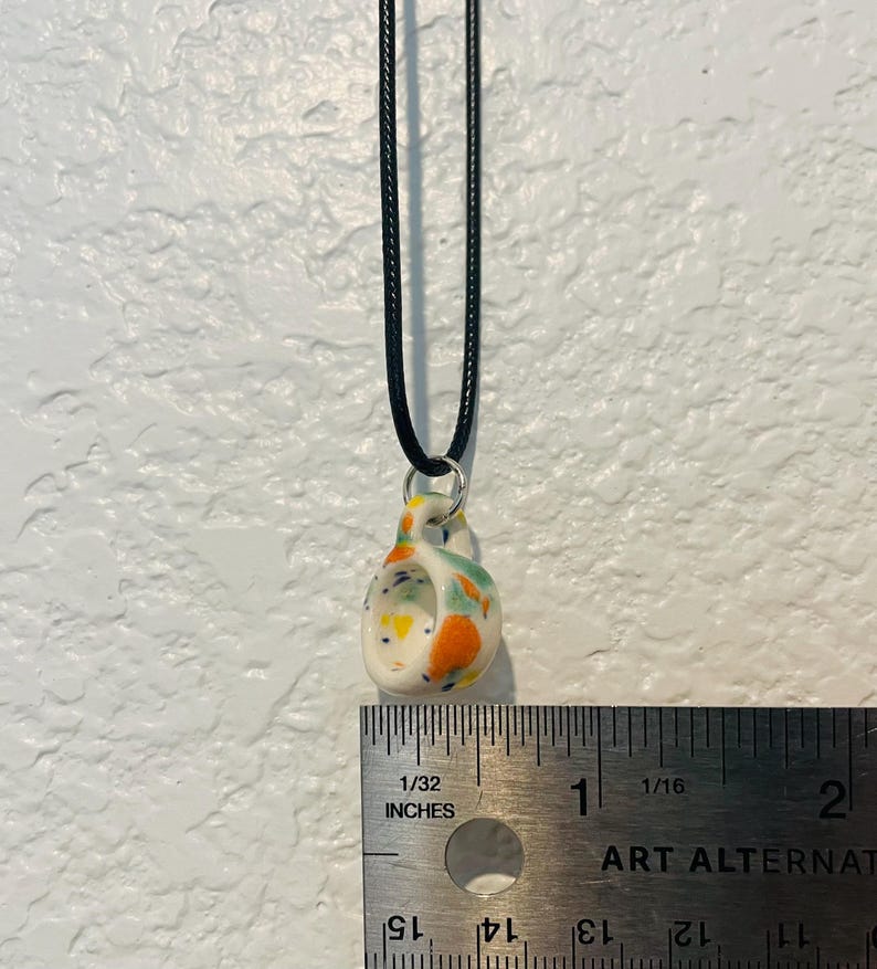 May include: A small, speckled ceramic pendant necklace with a black cord. The pendant is white with orange, green, and yellow accents. The pendant measures approximately 0.75 inches in height.
