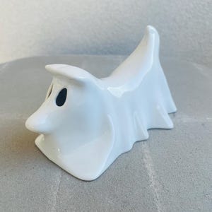 May include: A white ceramic ghost figurine with black eye holes. The ghost has a draped sheet design and a small, pointed nose. The figurine is a decorative item, likely for Halloween or spooky-themed decor.