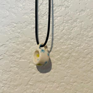 May include: A small, ceramic pendant necklace. The pendant is a miniature mug shape, white with blue and yellow accents. It hangs from a black cord. The background is a textured white wall.