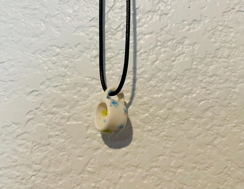 May include: A small, ceramic pendant necklace. The pendant is a miniature mug shape, white with blue and yellow accents. It hangs from a black cord. The background is a textured white wall.