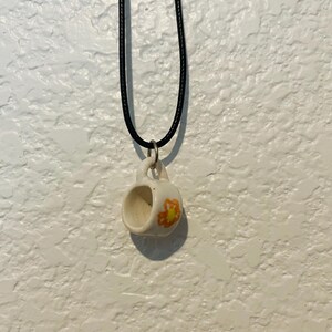 May include: A small, white ceramic mug pendant with a black cord necklace. The mug has a yellow and orange floral design on the side. The pendant is hanging against a textured white wall.
