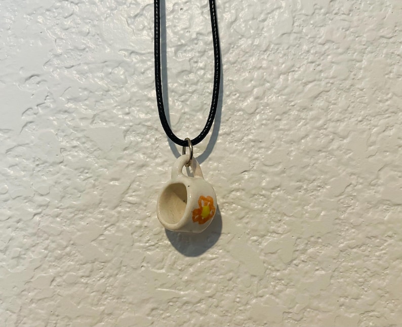May include: A small, white ceramic mug pendant with a black cord necklace. The mug has a yellow and orange floral design on the side. The pendant is hanging against a textured white wall.