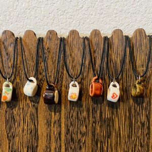 May include: A display of ceramic mug pendant necklaces on black cords. The miniature mugs are in various colors and designs, including white, orange, brown, and yellow. They are displayed on a wooden stand.