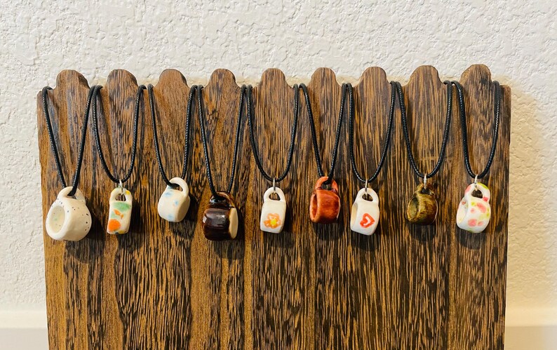 May include: A display of ceramic mug pendant necklaces on black cords. The miniature mugs are in various colors and designs, including white, orange, brown, and yellow. They are displayed on a wooden stand.