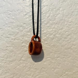 May include: A small, brown ceramic mug pendant hangs from a black cord. The pendant is a miniature replica of a coffee mug, with a handle and a hollow center. The pendant is suspended against a white textured wall.