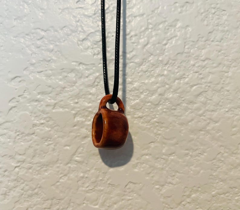 May include: A small, brown ceramic mug pendant hangs from a black cord. The pendant is a miniature replica of a coffee mug, with a handle and a hollow center. The pendant is suspended against a white textured wall.