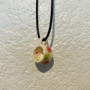 May include: A small, white ceramic teacup pendant with a black cord. The cup is decorated with pink and green spots. The pendant is suspended against a textured white wall.