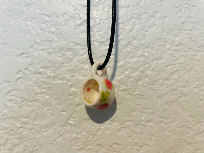 May include: A small, white ceramic teacup pendant with a black cord. The cup is decorated with pink and green spots. The pendant is suspended against a textured white wall.