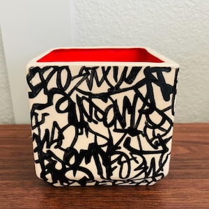 May include: A square ceramic planter with a cream-colored exterior featuring a dense, black hand-painted graffiti-style pattern of overlapping lines and abstract shapes. The interior of the vessel is finished in a vibrant, solid red glaze.