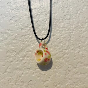 May include: A ceramic pendant necklace featuring a small, open-mouthed cup design. The cup is white with a pattern of red and green floral accents. The pendant hangs from a black cord necklace.