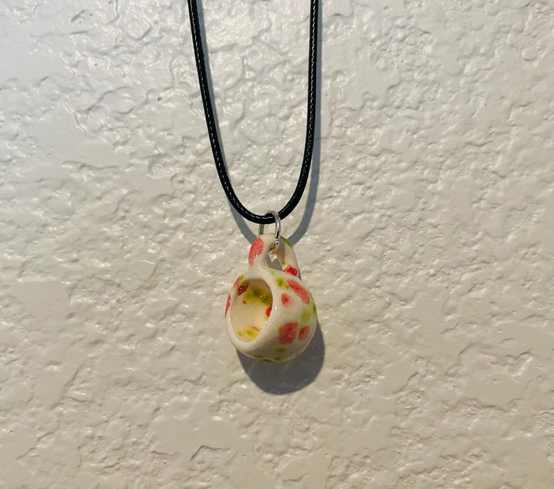 May include: A ceramic pendant necklace featuring a small, open-mouthed cup design. The cup is white with a pattern of red and green floral accents. The pendant hangs from a black cord necklace.