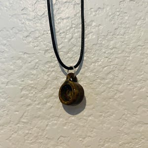 May include: A handmade ceramic pendant necklace. The pendant is a small, brown, cup-shaped charm with a dark interior. It hangs from a black cord necklace. The pendant is attached to the cord with a silver-colored ring.