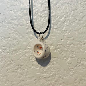 May include: A white ceramic pendant necklace shaped like a miniature mug. The mug is speckled with blue, orange, and red dots. It hangs from a black cord necklace with a silver-colored metal ring.