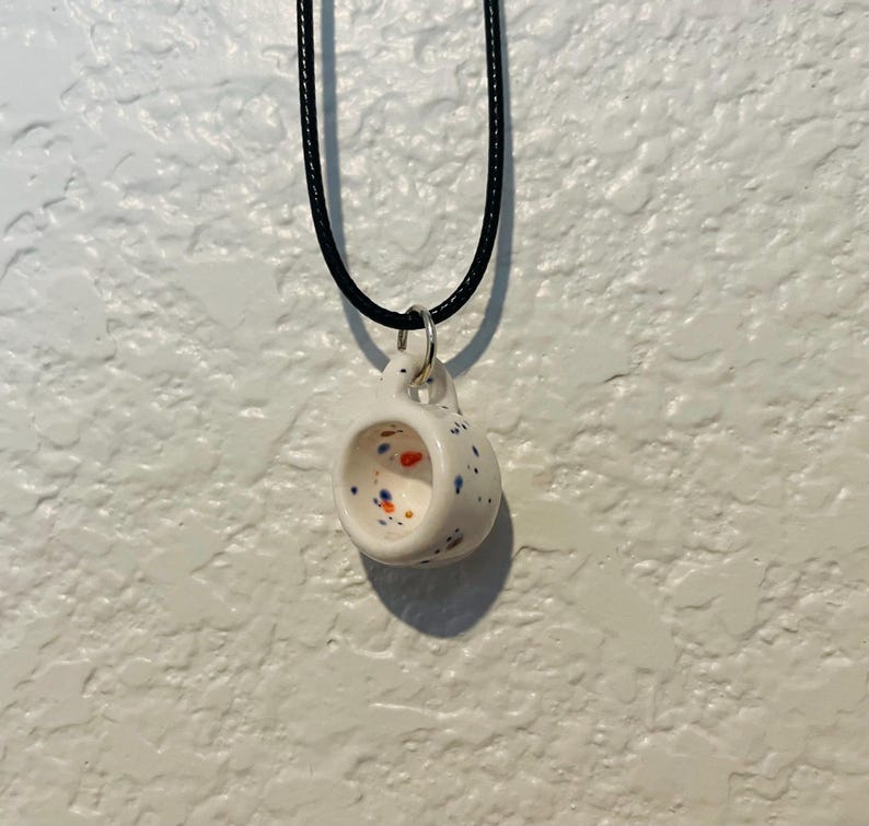 May include: A white ceramic pendant necklace shaped like a miniature mug. The mug is speckled with blue, orange, and red dots. It hangs from a black cord necklace with a silver-colored metal ring.